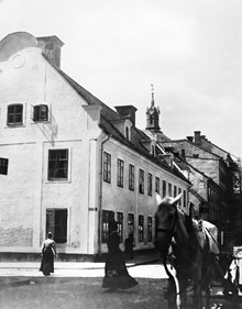 Några kvinnor och en häst passerar Storgatan 12 vid hörnet av Skeppargatan (t.v.). Hedvig Eleonora kyrkas torn i fonden