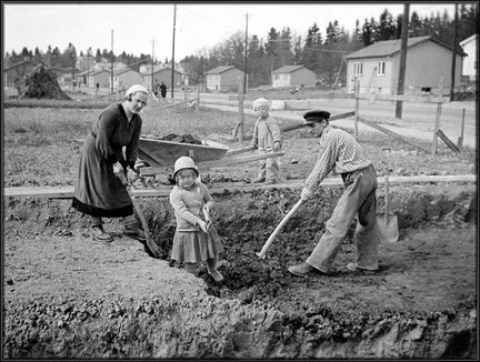 En familj, föräldrar och två små barn,  hjälps åt med att gräva med spadar och skottkärra för grunden till ett egnahemshus.