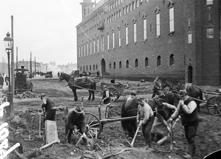 Svart-vit bild med Stadshusets fasad i bakgrunden. I förgrunden ser man ett tiotal arbetare som gräver i jorden och lassar kärror som dras av hästar.