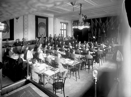Kostymklädda män sitter i rader vid bord med skriv- och ritmaterial. De är fotograferade bakifrån och är vända mot en estrad där en man står vid en talarstol.