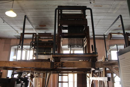 En jacquardmekanik i trä och järn ovanpå en vävstol i trä. På vävstolen är skrivet S:t E. Mekaniken är inte inkopplad. Det är en fabriksmiljö, väggarna är rosa och taket ljusgrått.