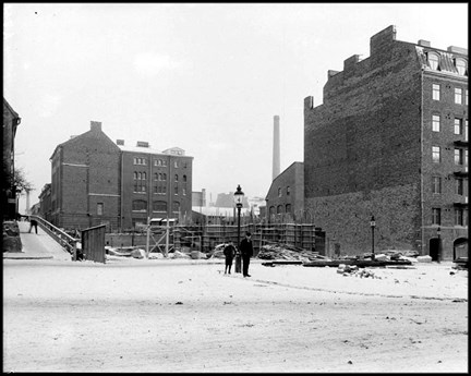 Gatubild med flerfamiljshus på Kungsholmstorg. Hörnhuset i kvarteret har grundlagts. En man och en pojke står framför huset. Det är vinter och snö på marken.