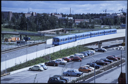 Ett blått tunnelbanetåg vid en parkering där det står bilar år 1977.