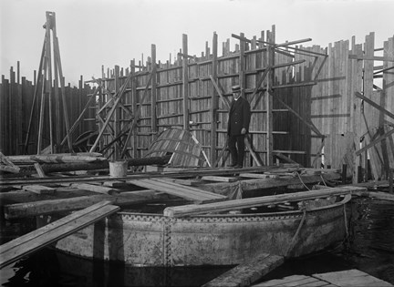 En arbetsplats med plank i bakgrunden och en stor cirkelrund form i förgrunden. Över formen går en man i hatt och kostym över några plankor som ligger utlagda.