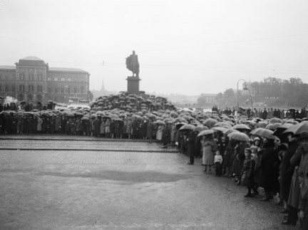 Folksamling vid Slottsbacken på Skeppsbron i väntan på begravningståget med stoftet efter de förolyckade från Andréexpeditionen. I fonden Nationalmuseet och Gustaf III:s staty.