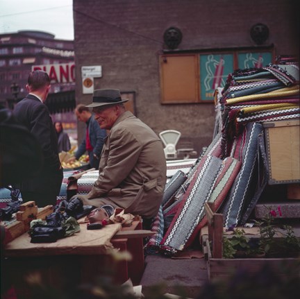 En man säljer plastmattor och figurer från ett stånd på Hötorget. I bakgrunden syns Konserthuset och en fruktförsäljare.