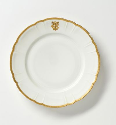 En vit tallrik i porslin med förgyllda kanter dekorerat med ett gyllene emblem.