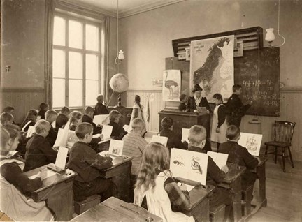 Klassrum med elever som sitter vid bänkarna. Framme vid katedern sitter en lärarinna framför en karta över Sverige-Norge