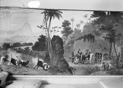En platsbyggd soffa klädd i randigt tyg framför en vägg tapetserad med en fransk tapet med ett motiv från Brasilien. Palmer, människor till häst och till fots. till vänster i bild ett runt bord med en duk med fransar.