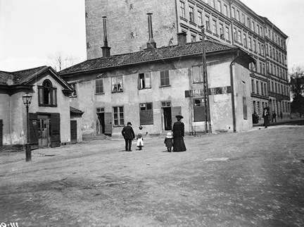 En äldre lägre byggnad och en högre i bakgrunden. På gårdsplanen framför husen syns en man och en kvinna med varsitt barn i handen.