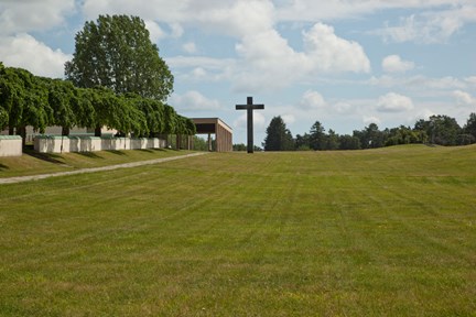 Ett stor öppet gräsfält. i bakgrunden står ett stort svart kors mot en blå himmel med vita molntussar. Till vänster träd och  en byggnad med pelare som innehåller kapell och ett krematorium.