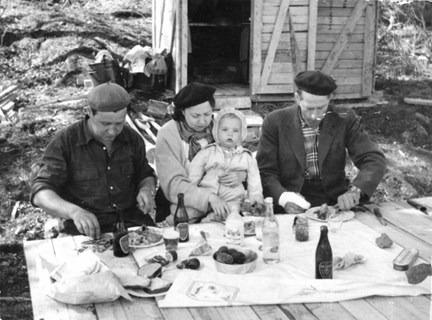 Två män, en kvinna och ett litet barn äter lunch tillsammans utomhus. I bakgrunden syns en liten träbod.