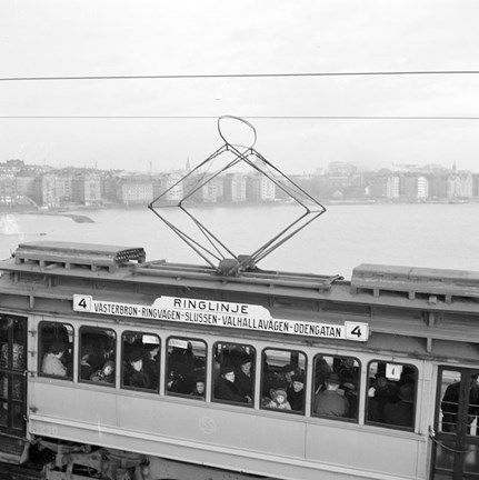 Spårvagn nummer 4 med texten "Ringlinje" på fasaden, fotograferad i svart-vitt snett ovanifrån. Flera människor syns i vagnen och i bakgrunden syns utsikten från Västerbron, mot Norr Mälarstrand.