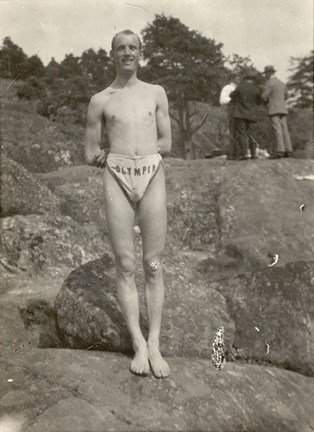 Foto på ung man i badbyxor med texten Olympia , stående på klippa.