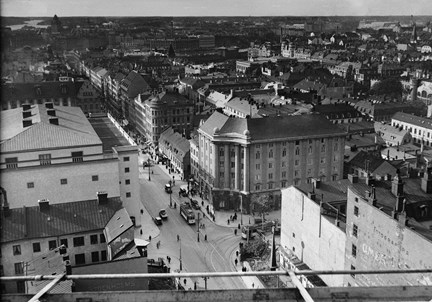 Utsikten från en hög byggnad. Nere på marken syns en större vägkorsning omgärdad av större hus, varv det ena är Stockholms konserthus och det andra en butiks- och kontorsfastighet. I korsningen rullar två spårvagnar och ett par bilar. I bakgrunden breder staden ut sig med en mängd större stenhus. 