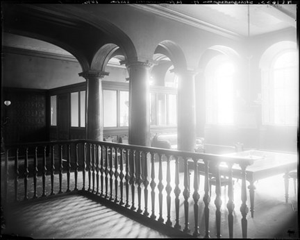 En sal med flera pelare och ett räcke av formsvarvat trä. Ljuset flödar in. I mitten står ett större bord och några stolar.