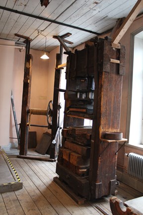 En stor press som går från golv till tak står framför två fönster. Bakom pressen syns ett rep och en vev som används för att spänna åt den. Väggarna är rosa.
