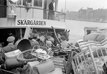 Fullastad skärgårdsbåt under midsommar med människor, bagage och en häst. Vy mot Skeppsbron och Södermalm