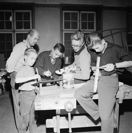 Fyra pojkar står kring och sitter på ett arbetsbord med sina slöjdarbeten. En äldre man ser på i bakgrunden.