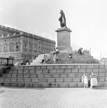 Skeppsbron, Gustav III:s staty. Sittande människor på trappstegen. I bakgrunden Stockholms slott