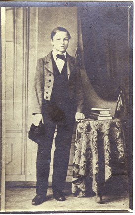 Svartvitt fotografi på Strindberg 13 år gammal, stående med mössa i handen vid ett bord och ett draperi .