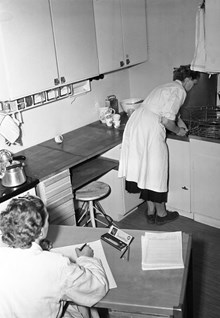 Interiör från kök på Rålambsvägen i Marieberg. En kvinna från Hemmens forskningsinstitut gör tidsstudier, med klocka och anteckningar, på en kvinna som arbetar vid köksbänken i köket.