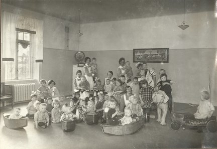 Svartvit bild av barn och sköterskor som är samlade för fotografering. Många av barnen sitter i gungleksaker eller har andra leksaker i händerna.