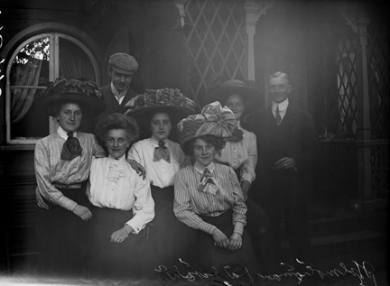 Fem kvinnor och två män sitter framför ett lusthus. Kvinnorna har stora hattar och blusar med knutna rosetter i halsen, männen har kavaj och slips. den ena mannen har en keps på huvudet. Alla tittar i kameran och ser glada ut. 