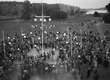 En dansbana med belysning är uppbyggd i parken. Några par dansar och en stor samling människor står runtomkring.