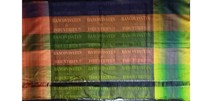 Ett sidentyg som är randigt i blått, rött och grönt. I de olika fälten syns en text där det står "BANCOVINSTEN åt INDUSTRIEN.!!"