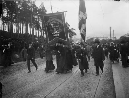 Svart-vit bild från en demonstration. I centrum syns tre kvinnor bära en fana med texten "Svenska Bryggeriarbetareförbundets avdelning 15". Kvinnorna är klädda i långa kjolar och runtom syns män i kostym och hatt.