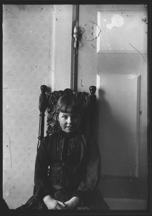 Greta Strindberg tio år gammal, iklädd en mörk klänning, sitter på en utsirad stol mot en vit vägg. Foto av pappa August Strindberg.