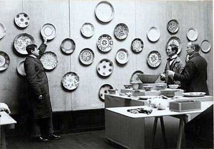Svartvitt fotografi av tre män som arbetar med skålar och fat i ett utställningsrum.