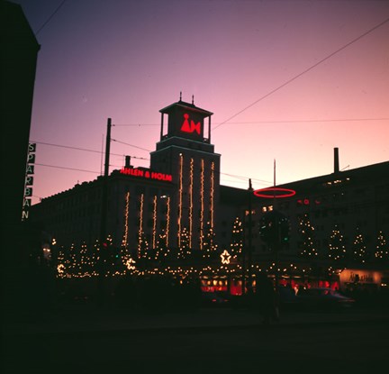 Färgbild tagen en mörk och sen vinterkväll. En varuhusbyggnad med ljusreklam och ljusslingor på fasaden. På taket står det "Åhlén och Holm" och "ÅH".