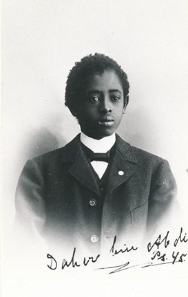 Svartvitt studioporträtt av en ung, svart man i vit skjorta och kostym. Nedtill ser man hans namnteckning.