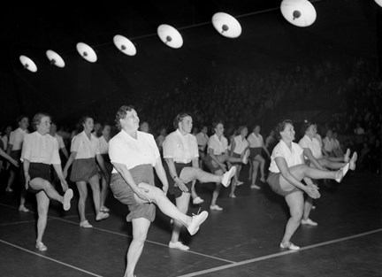 I en gymnastiksal, under en rad med runda lampor, syns kvinnor i gymnastikkläder utföra gymnastikrörelser. Kvinnorna är klädda i vita blusar och sportiga shorts.