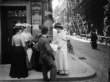 En kvinna i stor hatt och ljus klänning och kappa står i ett gathörn och delar ut tidningar till förbipasserande.