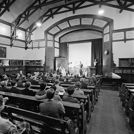I en stor sal med synliga träbjälkar i taket, sitter en mängd åhörare framför en scen och lyssnar på ett föredrag. 
