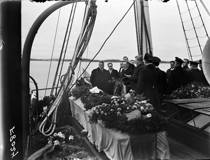 På ett fartygsdäck står tre kistor med svepning, dekorerade med blomsterkransar. Flera män lyssnar medan en man håller ett tal över de omkomna.