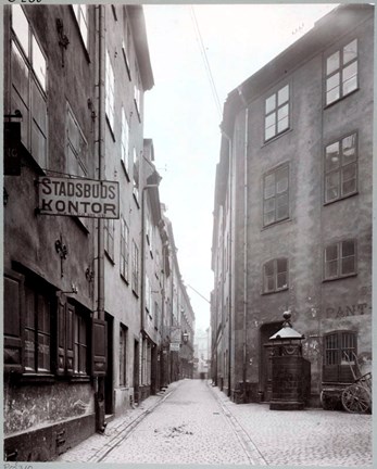 En öde gata, belagd med kullersten, omsluten av ålderdomliga stenhus. Ett stadsbudskontor och en pantbank syns, liksom en urinoar vid vad som ser ut som kanten av ett mindre torg.