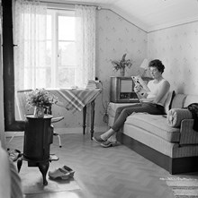 Vindsrum, Rävsnäs. Maud Lindgren läser veckotidning där rubriken ""Lycka"" skymtar. Järnkamin tillvänster