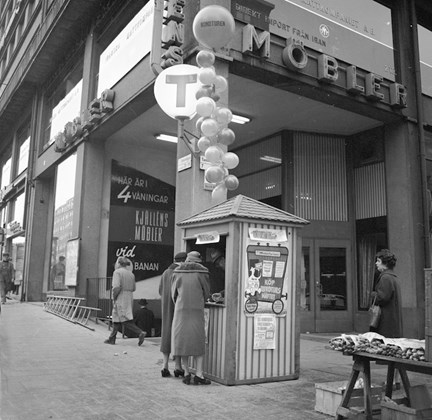 Två personer står framför luckan till en liten kiosk där massor av ballonger är fästa i taket. I bakgrunden ett varuhus.