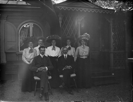 Ett lusthus med en veranda, täckt av växtlighet. Framför står fem stycken kvinnor i hattar och långa kjolar och blusar och två män sitter på varsin stol.