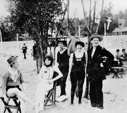 På en strand sitter en man och en kvinna på varsin pall. Intill den står två män och en kvinna. Mannen och kvinnan längst till höger ser glada och uppsluppna ur, de andra ler mer försiktigt. Alla är klädda i långbenta badkläder och bär hatt.