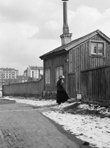 En kvinna står vid Holländarvägen 3. I fonden ses Götgatan 81 vid hörnet av Blekingegatan. Holländarvägen motsvarar ungefär nuv. Hallandsgatan (tidigare Skånegatan) och kv. Vågskivan