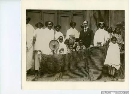 Hirsi Ige är på besök hos chefen på Etnografiska museet, Gerhard Lindblom, för att skänka honom en sköld, en dolk, en matta och en korgflaska. Kring dem står den somaliska gruppen, barn och vuxna i traditionella kläder.