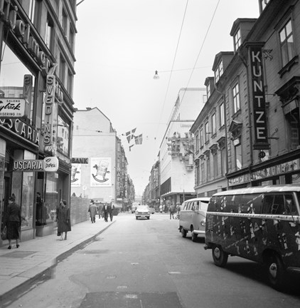 På gatan kör Volkswagen-bussar och den kantas av butiker. På skyltar står bland annat: Hjalmar Svedin. Oscaria. Bank. Kuntze.