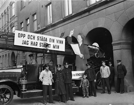 Stor pappersskylt med skådespelaren Adolf Jahr och texten "Opp och städa din vinn jag städar min" på en brandbil framför Johannes brandstation.