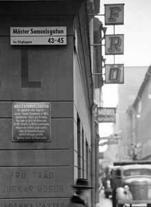 Gatuskylten vid Mäster Samuelsgatan 45 i hörnet av Beridarbansgatan söderut. En skylt berättar om Mäster Samuelsgatan. En butiksskylt tillhör Nordiska Fröhandeln AB