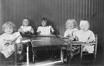 Fem små barn sitter vid bord på låga barnstolar i Engelbrekts barnkrubba, Valhallavägen 9.